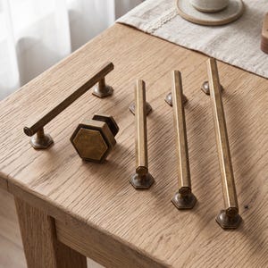 Puede incluir: Una colección de herrajes de latón antiguo que incluye cuatro tiradores de cajón alargados y un pomo de armario hexagonal. Cada pieza presenta un acabado metálico desgastado con un perfil hexagonal distintivo y bases de montaje hexagonales.