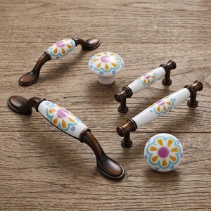May include: A collection of cabinet hardware including two arched handles, two straight bar handles, and two round knobs. Each piece features a white ceramic center with a painted floral pattern in yellow, light blue, and purple, set in bronze metal.