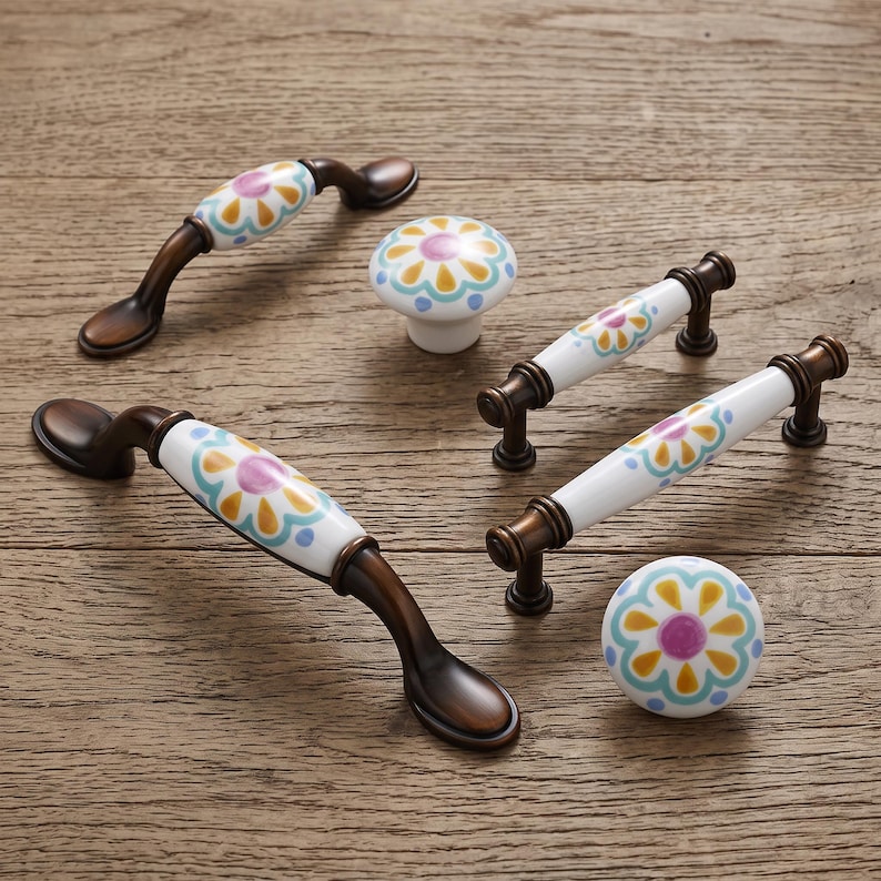 May include: A collection of cabinet hardware including two arched handles, two straight bar handles, and two round knobs. Each piece features a white ceramic center with a painted floral pattern in yellow, light blue, and purple, set in bronze metal.