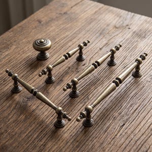 Può includere: Un set di ferramenta in ottone anticato composto da un pomello rotondo e quattro maniglie per cassetti allungate. Ogni pezzo presenta un design affusolato con basi di montaggio circolari a gradini e una finitura metallica patinata.