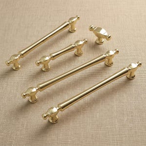 May include: A collection of four polished gold-tone metal cabinet hardware pieces, including three elongated drawer pulls with rounded ends and a single matching small knob, all featuring a smooth, reflective finish and classic cylindrical design.