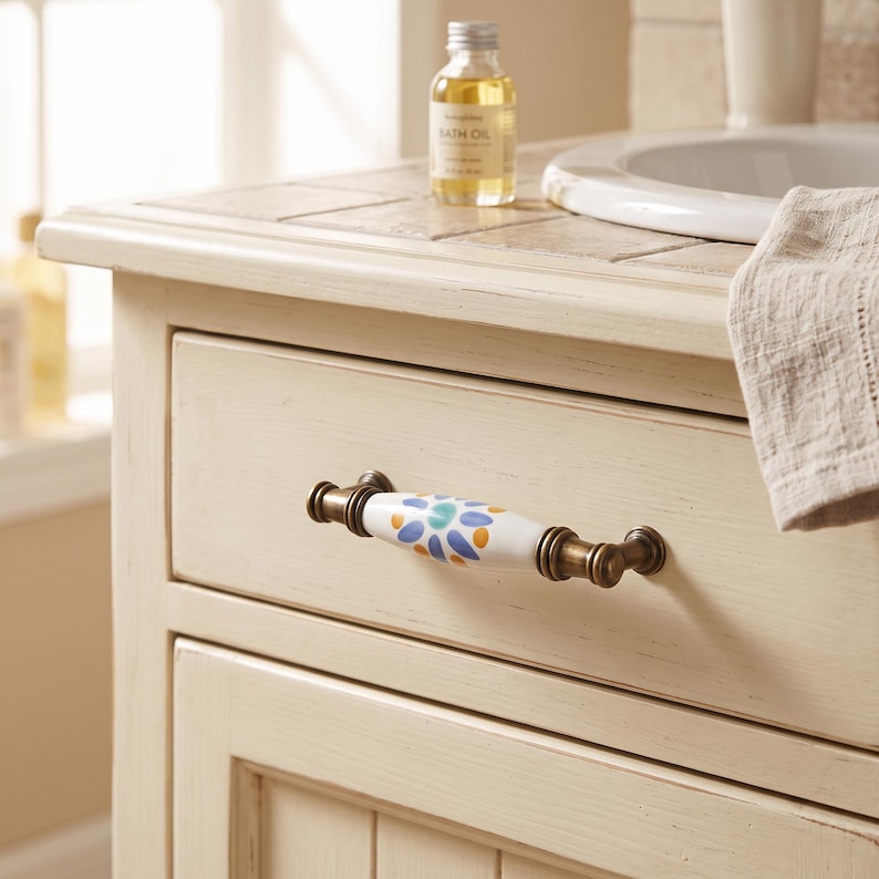 May include: A drawer pull featuring a white ceramic center with a blue, orange, and green floral pattern, flanked by antique brass-toned metal ends. The handle is mounted on a cream-colored wooden drawer front with a distressed, weathered finish.