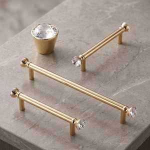 May include: A collection of gold-toned cabinet hardware including one conical knob and three cylindrical bar pulls. Each piece features a clear, faceted, diamond-like crystal accent at the ends or top, providing a reflective and metallic finish.