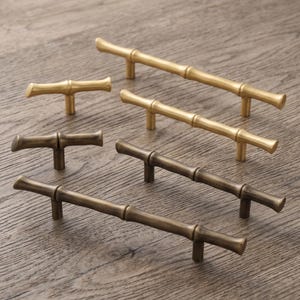 May include: A collection of six metal cabinet pulls and knobs featuring a bamboo-inspired design. The set includes three bright gold-toned pieces and three darker, antique bronze-toned pieces, each with a textured, segmented cylindrical shape.
