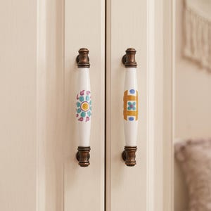 May include: Two vertical cabinet handles with white ceramic centers and dark bronze metal ends. The left handle features a teal and purple floral pattern, while the right handle displays a geometric design in mustard yellow and blue accents.