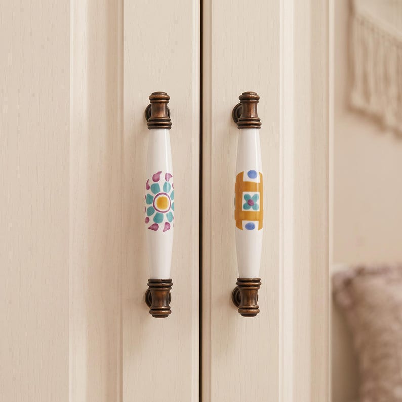 May include: Two vertical cabinet handles with white ceramic centers and dark bronze metal ends. The left handle features a teal and purple floral pattern, while the right handle displays a geometric design in mustard yellow and blue accents.