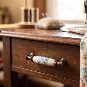 May include: A drawer pull featuring a white ceramic center with a blue and yellow floral pattern, attached to a dark brown wooden drawer with antique brass-toned metal hardware on each end of the handle, showing a smooth, rounded design.