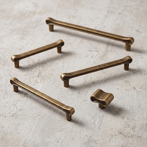 May include: A collection of five antique brass cabinet hardware pieces, including four elongated bar pulls and one single knob. Each piece features a distinct looped end design with a weathered, metallic finish and a smooth, curved profile.
