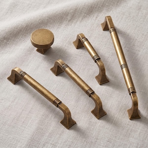 May include: A collection of antique brass hardware including one round knob and four drawer pulls of varying lengths. Each piece features a cylindrical bar design with ribbed detailing near the mounting points and angular, flared feet.