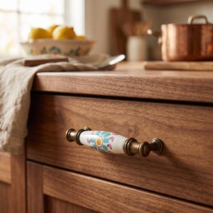 May include: A drawer pull featuring a white ceramic center with a painted teal and yellow floral pattern, flanked by antique brass metal hardware. The handle is mounted horizontally on a dark wood drawer front, showcasing a smooth finish.