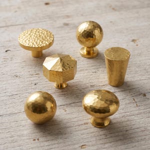 May include: A collection of six gold-toned metal cabinet knobs featuring various shapes including spherical, hexagonal, and flat-topped designs. Each knob displays a distinct hammered texture across its surface, providing a reflective finish.