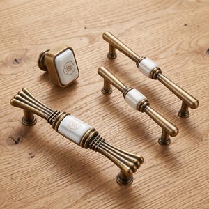 May include: A collection of cabinet hardware including one rectangular knob and three drawer pulls. Each piece features an antique brass finish combined with white ceramic inserts, some of which are decorated with a delicate gold floral line drawing.