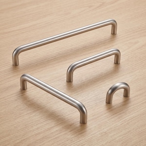 Puede incluir: Una colección de cuatro tiradores de armario de metal plateado cepillado de distintas longitudes, con un diseño liso y redondeado en forma de U con dos postes de montaje cilíndricos, excepto el pomo más pequeño en forma de arco.