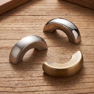 May include: Three semi-circular cabinet pulls in different metallic finishes. One pull has a polished chrome finish, one has a brushed silver finish, and one has a brushed brass finish. Each pull features a smooth, curved, arch-like shape.
