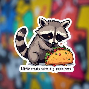 Op de afbeelding: Een sticker met een cartoon wasbeer die een taco eet. De wasbeer is grijs en wit met een zwart masker. De taco is gevuld met rode en groene ingrediënten. De tekst "Little treats solve big problems" staat eronder.