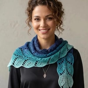 May include: A woman wearing a blue and teal crocheted shawl. The shawl has a leaf-like pattern along the edges and is draped around the neck and shoulders. A silver pendant hangs from a delicate chain.