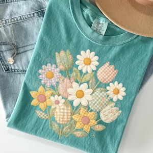 May include: A seafoam green t-shirt with a floral design featuring daisies and tulips, alongside light wash jeans, a tan hat, and a gold necklace. The word "seafom" is printed at the bottom right.
