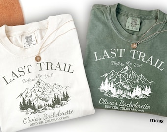 May include: Two crewneck t-shirts in ivory and moss green. Each shirt features a mountain and forest graphic with the text "LAST TRAIL Before the Veil", "Olivia's Bachelorette", and "DENVER, COLORADO 2026" printed in a contrasting colour.