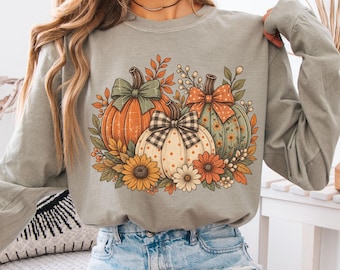 May include: A long-sleeve sandstone sweatshirt featuring a central graphic of three pumpkins adorned with bows, surrounded by autumn leaves and sunflowers. The pumpkins are orange, cream with polka dots, and sage green.