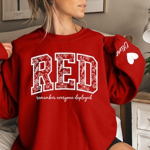 May include: A red sweatshirt with the word "RED" in white, floral-filled block letters. Below the word "RED" is the phrase "remember everyone deployed" in a script font. The sleeve has the name "Oliver" and a white heart.