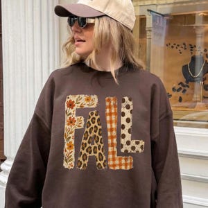 May include: A dark brown long-sleeve sweatshirt featuring the word FALL printed in large letters. Each letter has a distinct pattern: the F has a floral print, the A has a leopard print, the L has an orange gingham check, and the L has polka dots.