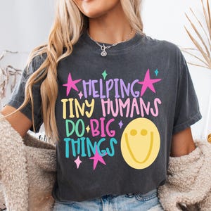 May include: A charcoal gray short-sleeve t-shirt featuring the text HELPING TINY HUMANS DO BIG THINGS in purple, yellow, pink, and blue lettering. The design includes pink stars, small blue and yellow diamond shapes, and a large yellow smiley face.