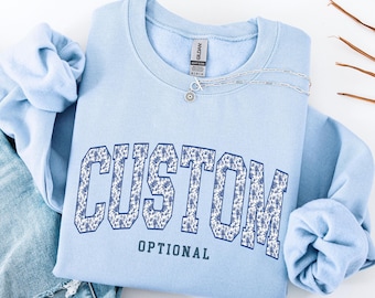 May include: A light blue crewneck sweatshirt featuring the word CUSTOM in large block letters with a blue and white floral pattern, and the word OPTIONAL in smaller blue text underneath. A silver necklace rests on the neckline of the garment.