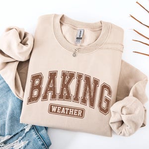 May include: A beige crewneck sweatshirt featuring the brown distressed text BAKING WEATHER in a collegiate font. The garment has long sleeves and a ribbed neckline. A silver-toned chain necklace with a small circular pendant rests on the collar.