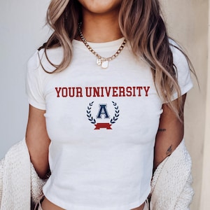 May include: White crop top featuring the text "YOUR UNIVERSITY" in red. Below the text is a navy blue letter "A" within a laurel wreath, with a red banner underneath. The shirt has short sleeves.
