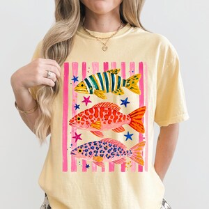 May include: Pale yellow t-shirt with a fish design. The graphic features three fish in yellow, red, and blue, set against a pink and white striped background. Blue and pink stars are scattered around the fish.