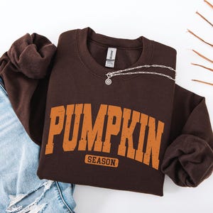 Peut inclure: Un sweat-shirt à col rond marron foncé avec le texte PUMPKIN SEASON imprimé dans une police universitaire orange vieillie. Le vêtement possède des manches longues et un col côtelé. Un collier à chaîne argentée avec un pendentif repose sur le col.