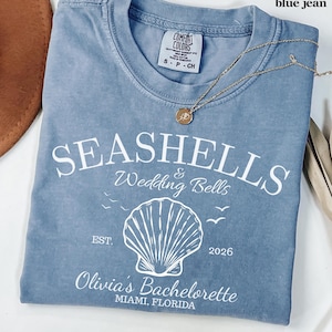 May include: A blue jean colored t-shirt featuring white screen-printed text that reads SEASHELLS & Wedding Bells, EST. 2026, and Olivia's Bachelorette MIAMI, FLORIDA, with a central seashell graphic and small bird silhouettes.
