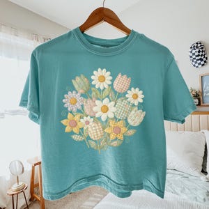 May include: A teal short-sleeve t-shirt featuring a central graphic of a bouquet of flowers. The floral design includes daisies and tulips in various pastel colours with gingham and plaid patterns, arranged in a circular cluster on the front.