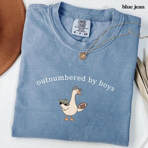 May include: Blue jean Comfort Colors t-shirt with the phrase "outnumbered by boys" and a cartoon goose wearing a hat and carrying a football and a toy truck. A gold necklace is draped across the shirt.