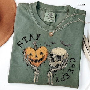 May include: A moss green crew neck t-shirt featuring a graphic print of two skeletal hands holding a heart-shaped jack-o'-lantern and a human skull. The text "STAY CREEPY" is printed in black around the graphic, accompanied by small bat silhouettes.