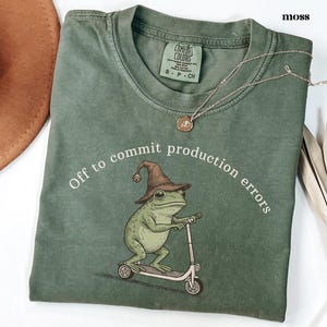 May include: A moss green t-shirt featuring a frog in a wizard hat on a scooter, with the text "Off to commit production errors". A silver necklace with a pendant is also visible. The t-shirt is a Comfort Colors brand.