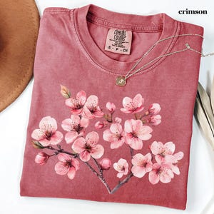 May include: Crimson-colored t-shirt with a floral design of pink cherry blossoms. The shirt has a crew neck and short sleeves. A silver necklace with a small pendant is draped on the shirt. The tag reads "Comfort Colors".