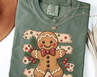 May include: A moss green crew neck t-shirt featuring a graphic of a smiling gingerbread person wearing a red and white polka dot bow tie. The design includes pine branches, red berries, and horizontal brush strokes with red and green dots.