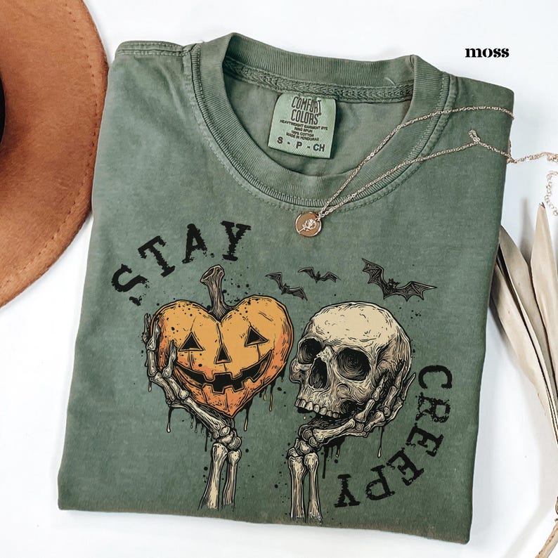 May include: A moss green crew neck t-shirt featuring a graphic print of two skeletal hands holding a heart-shaped jack-o'-lantern and a human skull. The text "STAY CREEPY" is printed in black around the graphic, accompanied by small bat silhouettes.