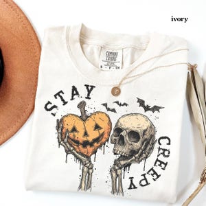 May include: An ivory t-shirt featuring a graphic print of two skeletal hands holding a heart-shaped jack-o'-lantern and a human skull. The text "STAY CREEPY" is printed in black lettering around the design, which also includes three small black bats.