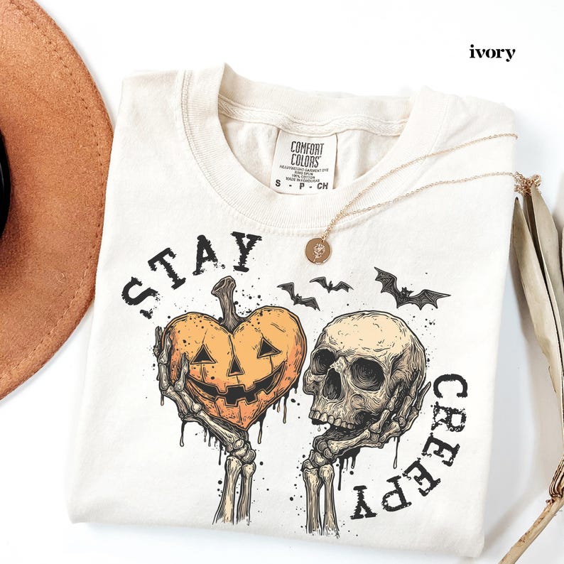May include: An ivory t-shirt featuring a graphic print of two skeletal hands holding a heart-shaped jack-o'-lantern and a human skull. The text "STAY CREEPY" is printed in black lettering around the design, which also includes three small black bats.