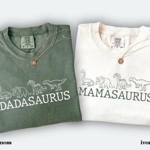 May include: Two t-shirts, one moss green and one ivory, each with a dinosaur design and text. The moss green shirt says "DADASAURUS" and the ivory shirt says "MAMASAURUS". Both shirts have a round gold pendant necklace.