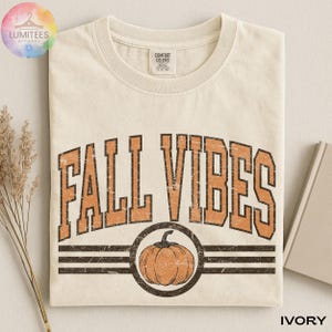 May include: An ivory crew neck t-shirt featuring the text FALL VIBES in a distressed orange font with a black outline. Below the text is a circular graphic containing an orange pumpkin illustration centered between two horizontal black lines.