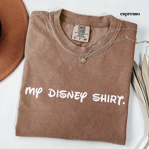 May include: Espresso-colored t-shirt with the text "my DISNEY SHIRT." A silver necklace with a pendant is draped across the shirt. The shirt has a crew neck and a tag that reads "COMFORT COLORS".