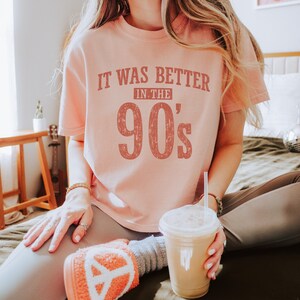 May include: A peach-colored short-sleeve t-shirt featuring the distressed dark red text IT WAS BETTER IN THE 90's printed on the front. The shirt has a crew neckline and a relaxed fit, showcasing a vintage-inspired graphic design on the chest.