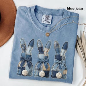 May include: A blue Comfort Colors t-shirt with three bunny appliqués. The bunnies have patchwork denim ears and bows, with white pom-pom tails. A gold necklace with a pendant is draped across the shirt. The text "blue jean" is visible.