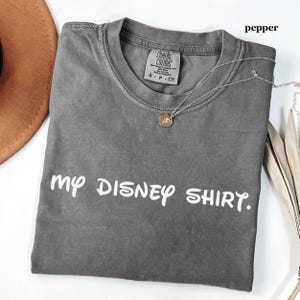 May include: A gray Comfort Colors t-shirt with the text "my DISNEY SHIRT." A silver necklace with a pendant rests on the shirt. The word "pepper" is in the upper right corner.
