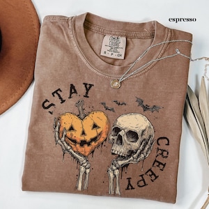 May include: An espresso-coloured t-shirt featuring a graphic print of two skeletal hands holding a heart-shaped jack-o'-lantern and a human skull. The text "STAY CREEPY" is printed in a distressed black font around the central illustration.