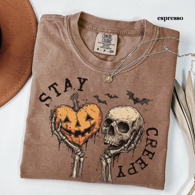 May include: An espresso-coloured t-shirt featuring a graphic print of two skeletal hands holding a heart-shaped jack-o'-lantern and a human skull. The text "STAY CREEPY" is printed in a distressed black font around the central illustration.