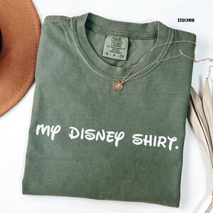 May include: A moss green t-shirt with the text "my DISNEY SHIRT." in white. A gold necklace with a pendant is on the shirt. The shirt is folded on a white surface.